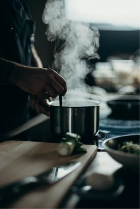 A person cooking in a kitchen with steam rising from a pot on the stove.