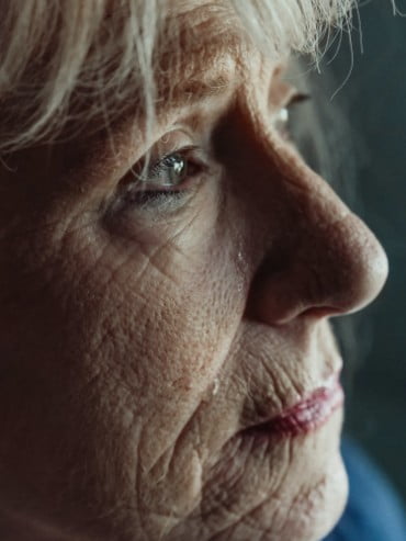 An elderly woman with wrinkles gazing off-camera. She has tears on her face.