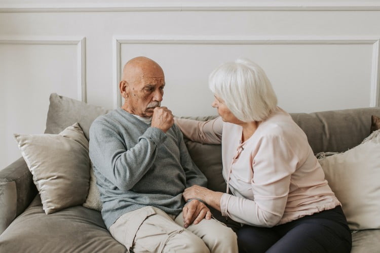 An elderly couple sat on a couch. The woman is comforting the man by putting her arm around his shoulder.